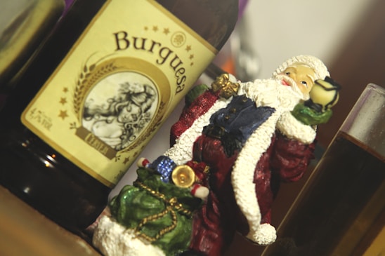 A Santa Claus figurine is positioned next to a beer bottle labeled 'Burguesa'. The figurine is holding a small cup and is adorned with traditional holiday colors, including red, green, and white, with hints of gold. A glass containing a dark liquid is partially visible in the background.
