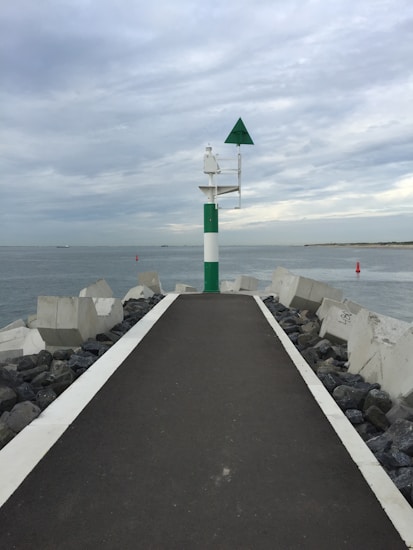 A paved path extends between large concrete blocks into a body of water. A navigational marker with a green and white pole topped with a green triangular sign stands at the end of the path. The sky is cloudy, and there are hints of distant land on the horizon.