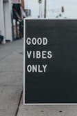A sidewalk sign features the words 'GOOD VIBES ONLY' in white letters on a black background. It is placed on a concrete sidewalk, with parts of buildings visible in the blurred background.