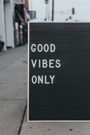 A sidewalk sign features the words 'GOOD VIBES ONLY' in white letters on a black background. It is placed on a concrete sidewalk, with parts of buildings visible in the blurred background.