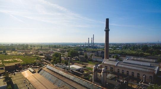 An expansive view of an industrial area with tall chimneys rising above large factory buildings. Green fields and scattered trees surround the complex, blending into the distant horizon. The sky is clear with a soft blue hue, indicating a sunny day.