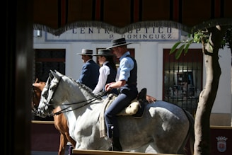 Three people dressed in traditional attire are riding horses through a sunny street. They are wearing wide-brimmed hats and dark vests. The backdrop includes a storefront with a sign that appears to be optician-related and some potted plants nearby.