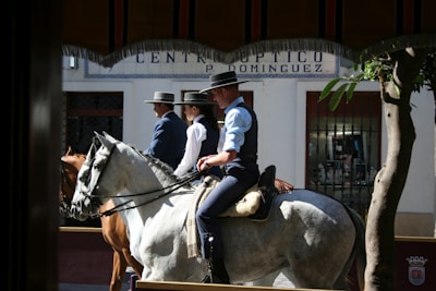Three people dressed in traditional attire are riding horses through a sunny street. They are wearing wide-brimmed hats and dark vests. The backdrop includes a storefront with a sign that appears to be optician-related and some potted plants nearby.