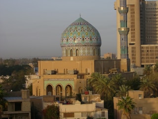 A historic mosque with an intricately designed blue and green mosaic dome and tall minaret surrounded by palm trees and urban buildings. The architecture is a blend of traditional and modern styles, with detailed geometric patterns in the tile work.