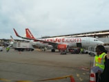 An easyJet airplane is parked on the tarmac, surrounded by ground service vehicles and personnel. The airplane is white with orange accents, and the easyJet logo is prominently displayed. Several workers in high-visibility vests are seen walking and organizing luggage and cargo near the aircraft. An additional service vehicle and cones are visible in the foreground.