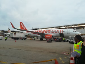 An easyJet airplane is parked on the tarmac, surrounded by ground service vehicles and personnel. The airplane is white with orange accents, and the easyJet logo is prominently displayed. Several workers in high-visibility vests are seen walking and organizing luggage and cargo near the aircraft. An additional service vehicle and cones are visible in the foreground.