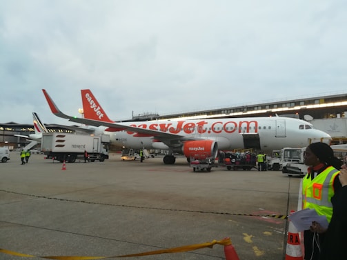 An easyJet airplane is parked on the tarmac, surrounded by ground service vehicles and personnel. The airplane is white with orange accents, and the easyJet logo is prominently displayed. Several workers in high-visibility vests are seen walking and organizing luggage and cargo near the aircraft. An additional service vehicle and cones are visible in the foreground.