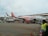 An easyJet airplane is parked on the tarmac, surrounded by ground service vehicles and personnel. The airplane is white with orange accents, and the easyJet logo is prominently displayed. Several workers in high-visibility vests are seen walking and organizing luggage and cargo near the aircraft. An additional service vehicle and cones are visible in the foreground.