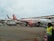 An easyJet airplane is parked on the tarmac, surrounded by ground service vehicles and personnel. The airplane is white with orange accents, and the easyJet logo is prominently displayed. Several workers in high-visibility vests are seen walking and organizing luggage and cargo near the aircraft. An additional service vehicle and cones are visible in the foreground.
