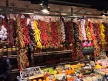 A vibrant market display features various dried peppers, garlic, and other spices hanging from the ceiling. The assortment includes red, yellow, and green peppers, with clusters of garlic adding to the variety. Below the hanging spices, fresh fruits and packaged spices are neatly arranged on the counter.