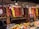 A vibrant market display features various dried peppers, garlic, and other spices hanging from the ceiling. The assortment includes red, yellow, and green peppers, with clusters of garlic adding to the variety. Below the hanging spices, fresh fruits and packaged spices are neatly arranged on the counter.