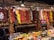 A vibrant market display features various dried peppers, garlic, and other spices hanging from the ceiling. The assortment includes red, yellow, and green peppers, with clusters of garlic adding to the variety. Below the hanging spices, fresh fruits and packaged spices are neatly arranged on the counter.