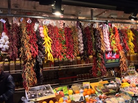 A vibrant market display features various dried peppers, garlic, and other spices hanging from the ceiling. The assortment includes red, yellow, and green peppers, with clusters of garlic adding to the variety. Below the hanging spices, fresh fruits and packaged spices are neatly arranged on the counter.