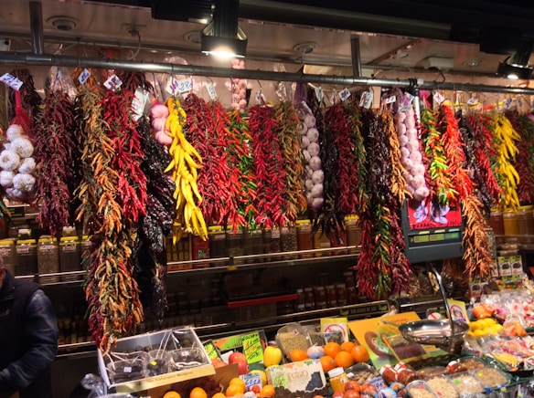 A vibrant market display features various dried peppers, garlic, and other spices hanging from the ceiling. The assortment includes red, yellow, and green peppers, with clusters of garlic adding to the variety. Below the hanging spices, fresh fruits and packaged spices are neatly arranged on the counter.