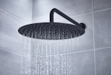 A large, round showerhead releasing streams of water in a tiled bathroom. The showerhead is mounted on an angled pipe extending from the wall, providing a gentle cascade of water.