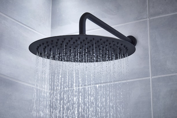 A large, round showerhead releasing streams of water in a tiled bathroom. The showerhead is mounted on an angled pipe extending from the wall, providing a gentle cascade of water.