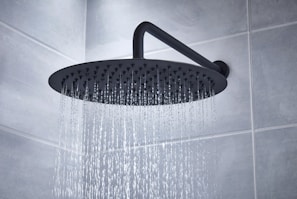 A large, round showerhead releasing streams of water in a tiled bathroom. The showerhead is mounted on an angled pipe extending from the wall, providing a gentle cascade of water.