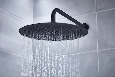 A large, round showerhead releasing streams of water in a tiled bathroom. The showerhead is mounted on an angled pipe extending from the wall, providing a gentle cascade of water.