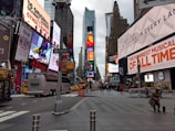 A busy urban street with numerous large, colorful billboards and advertisements on the buildings. The scene includes several buses and vehicles, as well as pedestrians walking on the sidewalks. Tall skyscrapers rise in the background, and a cloudy sky can be seen above.