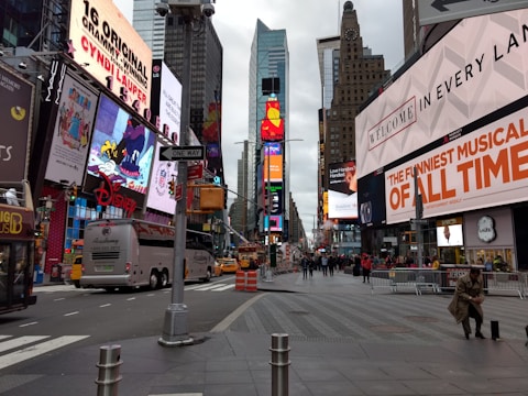 A busy urban street with numerous large, colorful billboards and advertisements on the buildings. The scene includes several buses and vehicles, as well as pedestrians walking on the sidewalks. Tall skyscrapers rise in the background, and a cloudy sky can be seen above.