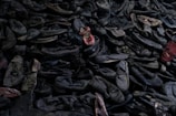 A large pile of old, worn-out shoes, mostly dark colored, with a few red ones standing out. The shoes appear to be randomly stacked, creating a chaotic and cluttered scene.