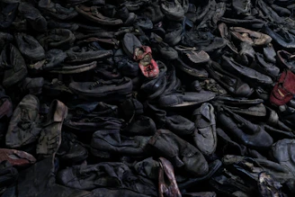A large pile of old, worn-out shoes, mostly dark colored, with a few red ones standing out. The shoes appear to be randomly stacked, creating a chaotic and cluttered scene.