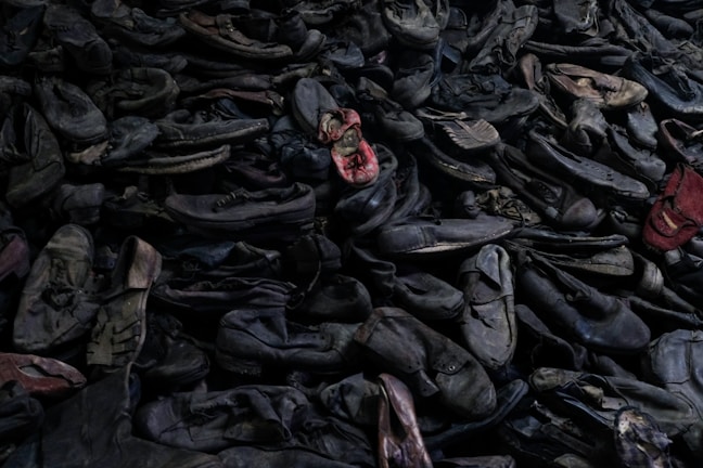 A large pile of old, worn-out shoes, mostly dark colored, with a few red ones standing out. The shoes appear to be randomly stacked, creating a chaotic and cluttered scene.