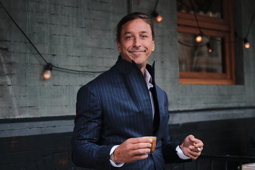 man wearing black suit holding brown mug near black string lights