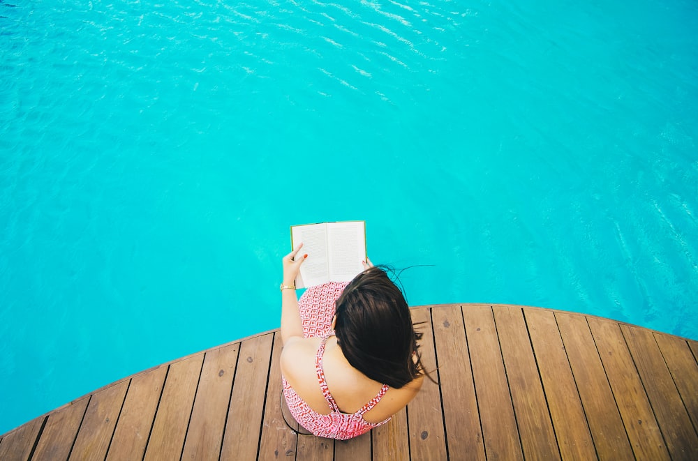 woman sitting on poolside dock while riding book