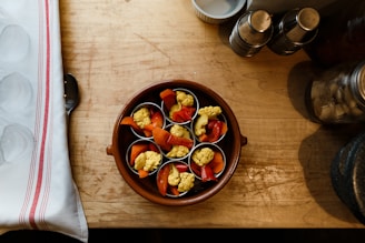 A wooden table holds a brown ceramic dish containing small cups filled with mixed vegetables, including yellow cauliflower and red peppers. To the left is a folded white cloth with red stripes, partially covering a metal spoon. In the top right corner, there are metal containers and a glass jar.