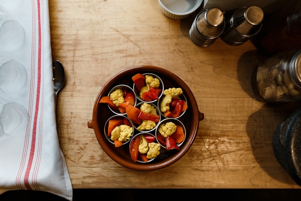 A wooden table holds a brown ceramic dish containing small cups filled with mixed vegetables, including yellow cauliflower and red peppers. To the left is a folded white cloth with red stripes, partially covering a metal spoon. In the top right corner, there are metal containers and a glass jar.