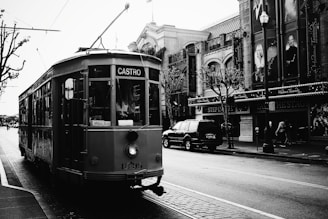 A historic streetcar marked with the destination 'Castro' is traveling along a cobblestone street lined with vintage buildings and leafless trees. The streetcar features a sign indicating 'Milan, Italy.' Adjacent to the streetcar, there is a large SUV parked in front of buildings with banners and signs, and a few pedestrians are visible. The image is in black and white, contributing to a nostalgic atmosphere.