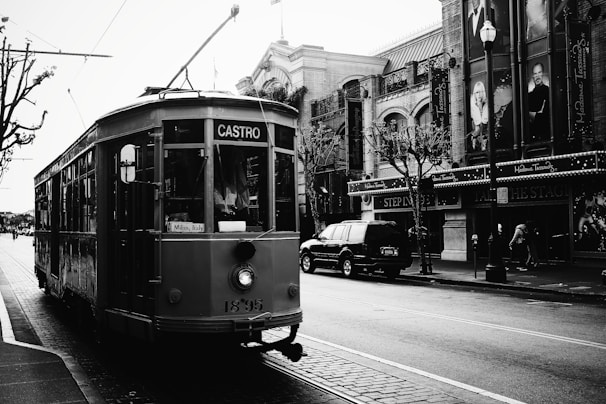 A historic streetcar marked with the destination 'Castro' is traveling along a cobblestone street lined with vintage buildings and leafless trees. The streetcar features a sign indicating 'Milan, Italy.' Adjacent to the streetcar, there is a large SUV parked in front of buildings with banners and signs, and a few pedestrians are visible. The image is in black and white, contributing to a nostalgic atmosphere.