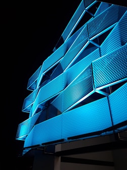 A building facade illuminated with vibrant blue lights, showcasing geometric patterns and a modern architectural design. The structure features repeating triangular shapes with textured surfaces that create a dynamic visual effect against the night sky.