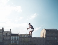 A person is performing a stunt on a scooter in mid-air against a backdrop of historic buildings under a partly cloudy sky.