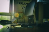 A dimly lit office space featuring a large computer monitor on a wooden desk, surrounded by various office supplies. Papers and cables are present, with a window to the left letting in natural light. Some motivational posters and books are visible in the background.