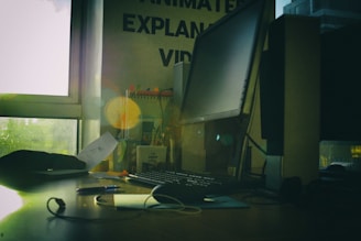 A dimly lit office space featuring a large computer monitor on a wooden desk, surrounded by various office supplies. Papers and cables are present, with a window to the left letting in natural light. Some motivational posters and books are visible in the background.