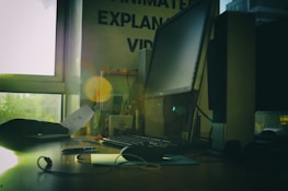 A dimly lit office space featuring a large computer monitor on a wooden desk, surrounded by various office supplies. Papers and cables are present, with a window to the left letting in natural light. Some motivational posters and books are visible in the background.