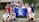A group of young people gathered together outdoors in front of a building, holding a blue flag with the Earth on it and the words 'PHS Global Ecology.' The group appears cheerful and diverse, standing closely together.