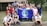 A group of young people gathered together outdoors in front of a building, holding a blue flag with the Earth on it and the words 'PHS Global Ecology.' The group appears cheerful and diverse, standing closely together.
