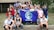 A group of young people gathered together outdoors in front of a building, holding a blue flag with the Earth on it and the words 'PHS Global Ecology.' The group appears cheerful and diverse, standing closely together.
