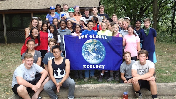 A group of young people gathered together outdoors in front of a building, holding a blue flag with the Earth on it and the words 'PHS Global Ecology.' The group appears cheerful and diverse, standing closely together.
