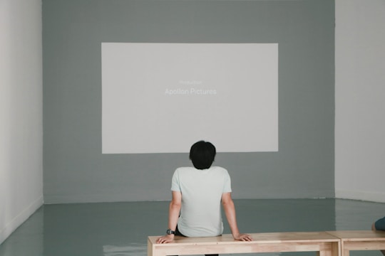 A person sits on a wooden bench facing a projected screen on a gray wall. The screen displays 'Production Apollon Pictures.' The room has minimalistic decor with a polished floor and white walls.
