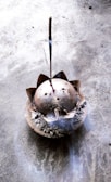 A lit incense stick is placed in an ash-filled, dome-shaped incense holder with a lotus petal design on a textured stone surface. Wisps of smoke gently rise from the incense, creating a subtle, calming atmosphere.