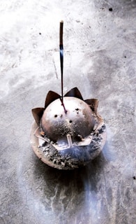 A lit incense stick is placed in an ash-filled, dome-shaped incense holder with a lotus petal design on a textured stone surface. Wisps of smoke gently rise from the incense, creating a subtle, calming atmosphere.