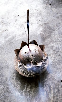 A lit incense stick is placed in an ash-filled, dome-shaped incense holder with a lotus petal design on a textured stone surface. Wisps of smoke gently rise from the incense, creating a subtle, calming atmosphere.