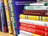 A collection of business and motivational books on a shelf, featuring titles like 'Greening Your Business,' 'Billions of Entrepreneurs,' 'Great Again,' and 'Higher Ambition.' The books are arranged vertically, showing a variety of colors and sizes.