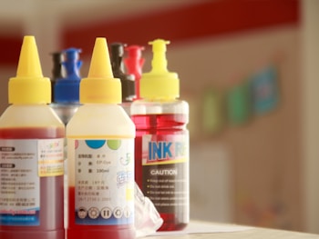 Several bottles with yellow caps are arranged closely together. The bottles have labels with text and colorful designs, and contain what appears to be ink or dye. The surroundings are blurred, suggesting a shallow depth of field.