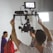 A person operates a camera rig with a mounted screen, capturing footage of a group of people in the background. The setting appears to be a studio or set, with professional lighting visible.