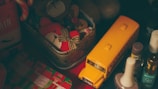 The image contains a collection of items including a yellow toy school bus, a tin containing stuffed toy bears, bottles of nail polish, and a festive gift box. The scene is dimly lit, creating a cozy, nostalgic atmosphere.